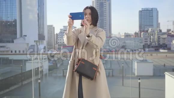 自信的黑发白种人女人站在城市街道上拍照的肖像使用智能手机的成年女士视频的预览图