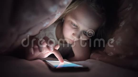 一个年轻的小女孩在睡觉后很晚躲在毯子下使用数字平板智能手机设备孤独视频的预览图