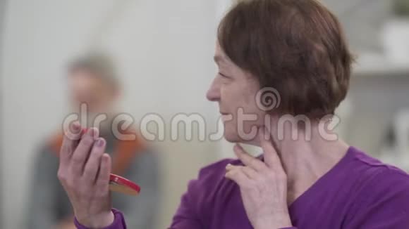 脸上挂着笑容的高加索女人看着手镜女性退休人员回头看模糊的男人和视频的预览图