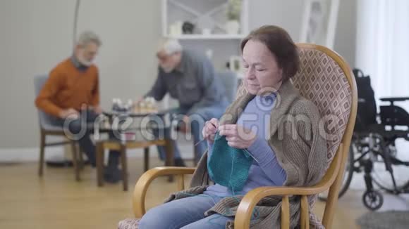 老白种人女人在扶手椅上编织就像模糊的男人在背景下下棋居住在视频的预览图
