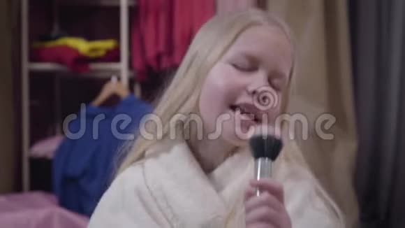 一个美丽的金发白种人女孩用化妆刷作为麦克风唱歌穿着白色浴袍的阳性儿童视频的预览图