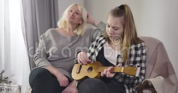 年轻漂亮的白种人女孩在室内为祖母演奏四弦琴和唱歌有才华的孙女和视频的预览图