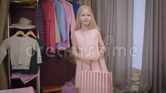 快乐的白种人金发女孩的肖像穿着粉红色的连衣裙站在家里带着购物袋快乐的孩子视频的预览图