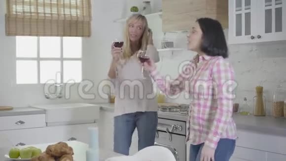 在现代厨房里拿着酒杯跳舞的成年白种人妇女的侧视两个朋友一起唱歌玩视频的预览图