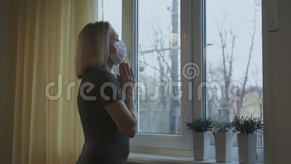 戴着医用面具的女人用双手在窗前祈祷视频的预览图