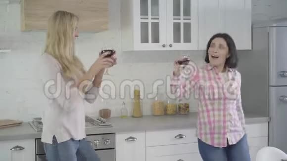 快乐的成年白种人妇女在现代厨房跳舞碰酒杯和喝酒的肖像两名女性视频的预览图
