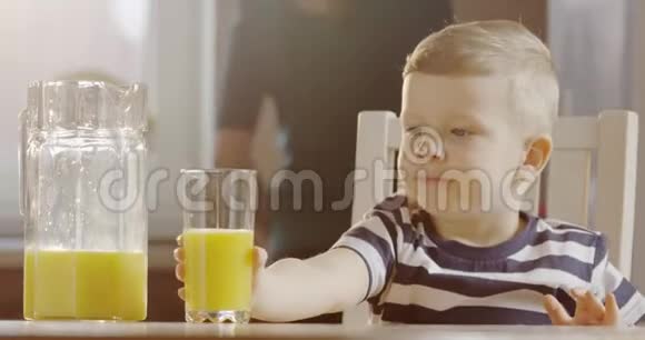 坐在桌子旁的小男孩喝着玻璃杯里的橙汁妈妈在厨房里用太阳照明弹做饭视频的预览图