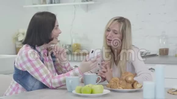 积极的黑发白种人女人向朋友展示新的智能手机快乐的女人坐在桌边聊天视频的预览图