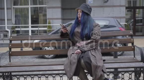 年轻美丽的高加索女人坐在城市街道的长凳上签名的肖像优雅的女士使用智能手机视频的预览图