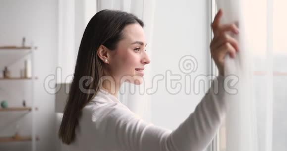 年轻的微笑女子打开窗帘透过窗户视频的预览图