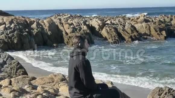 戴着耳机的男人在太平洋岩石海岸练习瑜伽带着壮丽的海浪视频的预览图