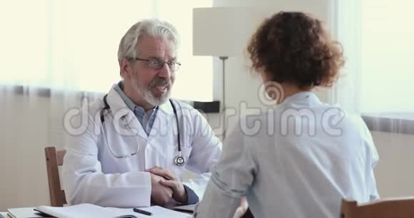 微笑老年男性医生咨询握手年轻女性患者视频的预览图