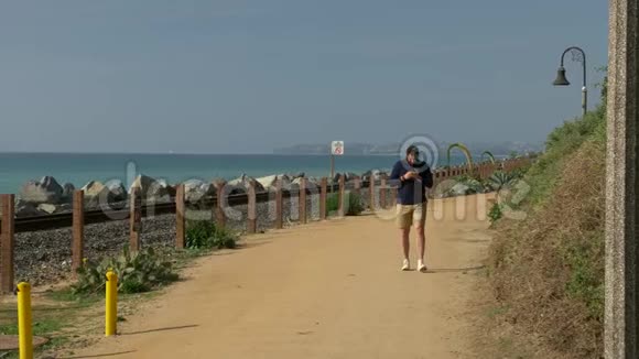 一个英俊的年轻人一个穿着海军蓝衬衫短裤和眼镜的男人正沿着加利福尼亚海岸圣克莱门特走着视频的预览图