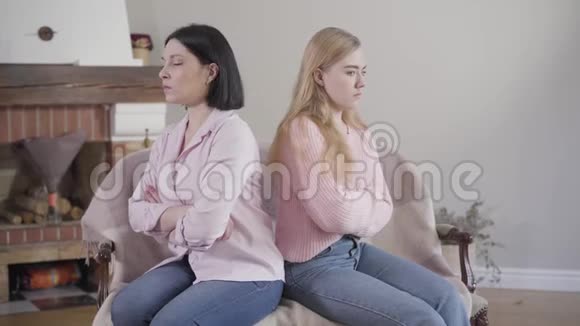 侧面观看成年白种人黑发女人和少女背靠背坐在沙发上母亲和女儿争论视频的预览图