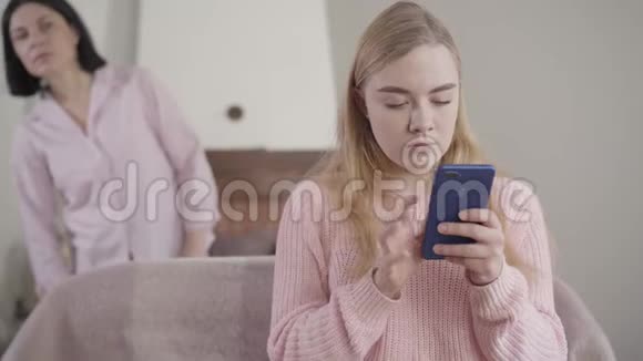 当她的母亲从后台看着屏幕时一台摄像机正在接近焦虑的高加索女孩在智能手机上打字视频的预览图