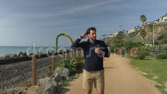 一个英俊的年轻人一个穿着海军蓝衬衫短裤和眼镜的男人正在沿着加利福尼亚海岸向前走视频的预览图