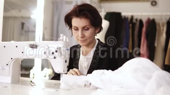 一位成年女裁缝的肖像在一台缝纫机上工作工作在一件白色手工制作的连衣裙上时尚服装视频的预览图