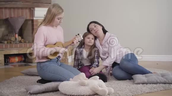 漂亮的金发白种人女孩在家为母亲和妹妹演奏四弦琴天才少年娱乐家庭视频的预览图
