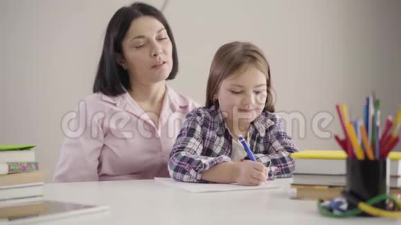 可爱的高加索女学生和妈妈坐在一起写在练习本上的肖像布鲁内特母亲帮助女儿视频的预览图