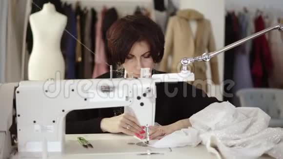 一位成年女裁缝的肖像在一台缝纫机上工作工作在一件白色手工制作的连衣裙上时尚服装和时装视频的预览图