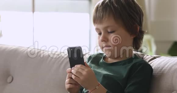 小可爱的男孩坐在沙发上在家里使用智能手机视频的预览图