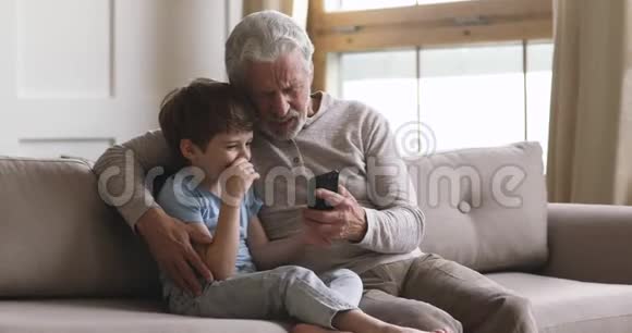 聪明的小男孩教年长的成熟祖父使用智能手机视频的预览图
