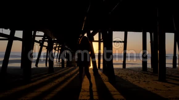 在美丽的橘红色夕阳下的码头下行走的女性剪影在壮观的阳光和太阳耀斑的木码头上视频的预览图