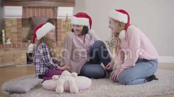 戴着圣诞帽坐在壁炉前的软地毯上笑着的快乐白人家庭的肖像母亲视频的预览图