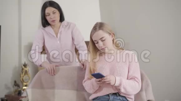漂亮的金发白种人女孩使用智能手机和听成人女人在后台大喊大叫的肖像青少年视频的预览图