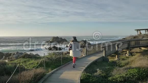 积极成熟的高白种人妇女穿着白色外套和温暖的帽子在太平洋海岸的桥上散步享受着视频的预览图