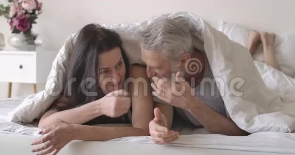 积极的高加索夫妇躺在毯子下的床上聊天和大笑微笑着成熟的男人视频的预览图