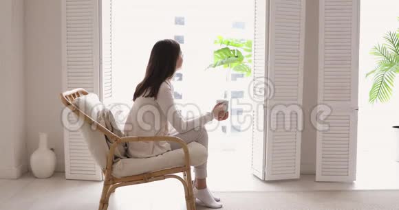 放松的年轻女子喝着茶透过窗户望着公寓视频的预览图
