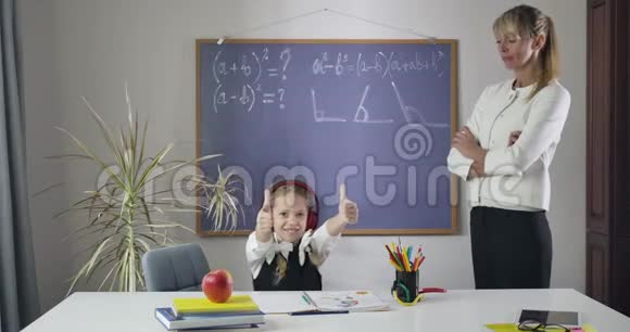 一个戴着耳机坐在餐桌前的高加索小女孩站在她旁边的成年家庭教师面前竖起大拇指视频的预览图