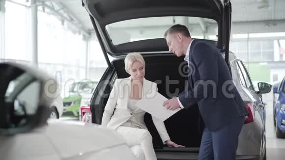 汽车经销商展示文件给年轻的金发白种人女人坐在汽车后备箱成功的商人视频的预览图