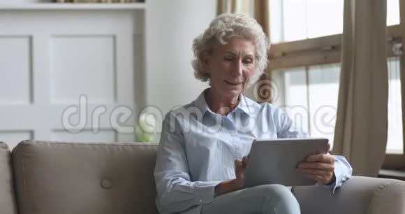 微笑的老年妇女坐在舒适的沙发上使用数字平板电脑视频的预览图