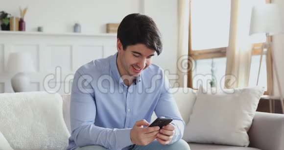 微笑的千禧一代用智能手机坐在沙发上视频的预览图