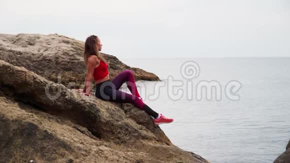 美丽的运动女郎穿着运动服坐在海边的岩石上背景是美丽的风景视频的预览图
