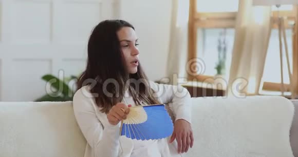 精疲力竭的年轻女士坐在沙发上挥舞着扇子视频的预览图