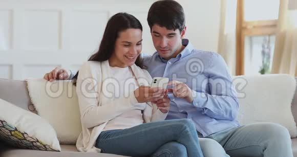 一对快乐的千禧夫妇用智能手机坐在沙发上聊天视频的预览图