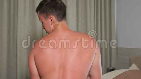 英俊的男人摸着被太阳晒伤的背部坐在卧室里感到疼痛视频的预览图