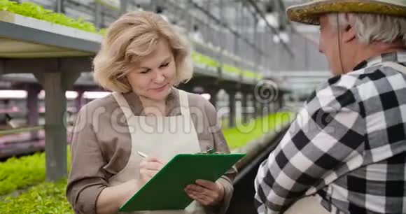 开朗的金发白种人女性灰色的眼睛在温室里听成熟的男性农学家写作视频的预览图