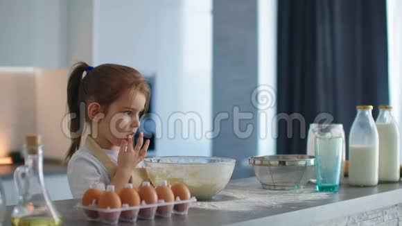 一个小女孩舔了舔她的手指站在厨房里围着围裙和她妈妈一起做面团视频的预览图