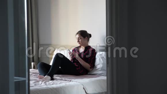 年轻女子坐在大床上卧室里有白色枕头和智能手机视频的预览图