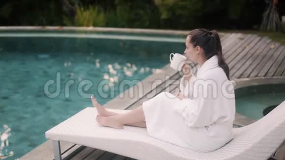 穿着浴袍带杯子的宁静女士坐在靠近游泳池的沙发上视频的预览图