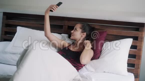 穿着天鹅绒睡衣的漂亮黑发女孩在床上自拍视频的预览图
