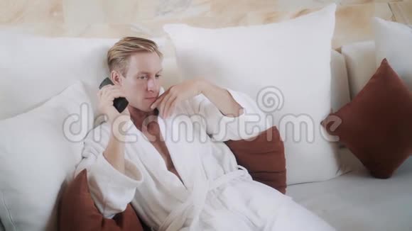 穿着浴袍的金发男人在舒适的床上打开电视视频的预览图