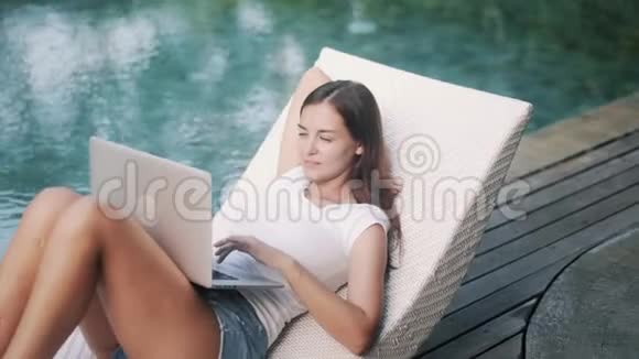 年轻女子在度假村游泳池附近的笔记本电脑上工作视频的预览图