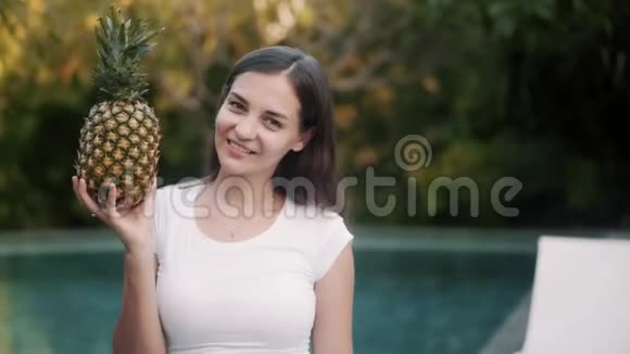 开朗的女人拿着成熟的菠萝对着游泳池视频的预览图