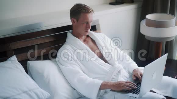 穿长袍的漂亮男人在舒适的床上工作笔记本电脑视频的预览图