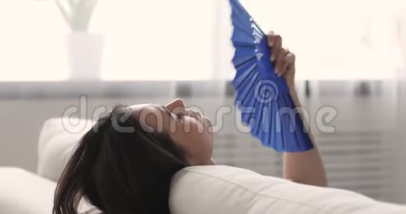 疲惫的女士挥舞着风扇在家里的沙发上降温视频的预览图